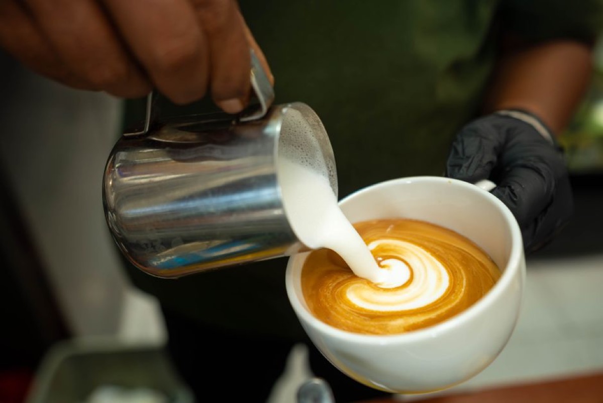 Close-up of barista mid-pour creating heart pattern in white espresso cup, milk stream visible